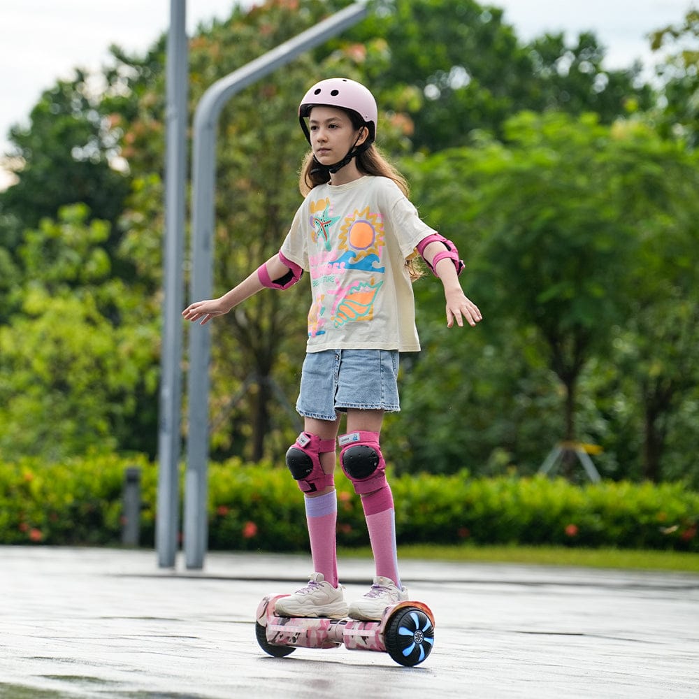 A young girl wearing a pink helmet and protective gear rides a hoverboard H2 in a park, balancing confidently. She sports a colorful t-shirt, denim shorts, and pink knee-high socks, with lush green trees in the background.