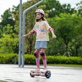 A young girl wearing a pink helmet and protective gear rides a hoverboard H2 in a park, balancing confidently. She sports a colorful t-shirt, denim shorts, and pink knee-high socks, with lush green trees in the background.