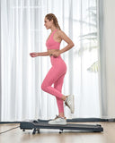 A woman in pink athletic wear is jogging on a treadmill in a bright room with sheer curtains and a glimpse of a plant outside.