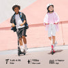Two children ride scooters ik1s on a sunny day, both wearing helmets for safety. The child on the left rides a black scooter, dressed in a black and white outfit, while the child on the right rides a pink scooter, wearing a pink jacket and white shorts. A pastel pink wall serves as the backdrop.