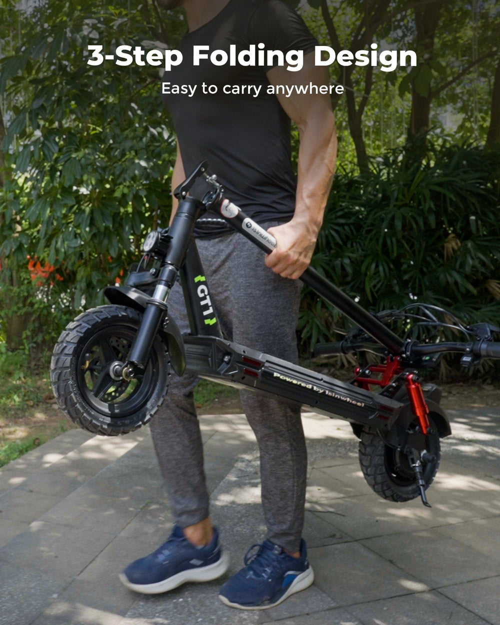 A person in casual athletic wear carries a folded black scooter GT1 with chunky tires along a paved path in a park setting, showcasing its 3-step folding design for easy portability.