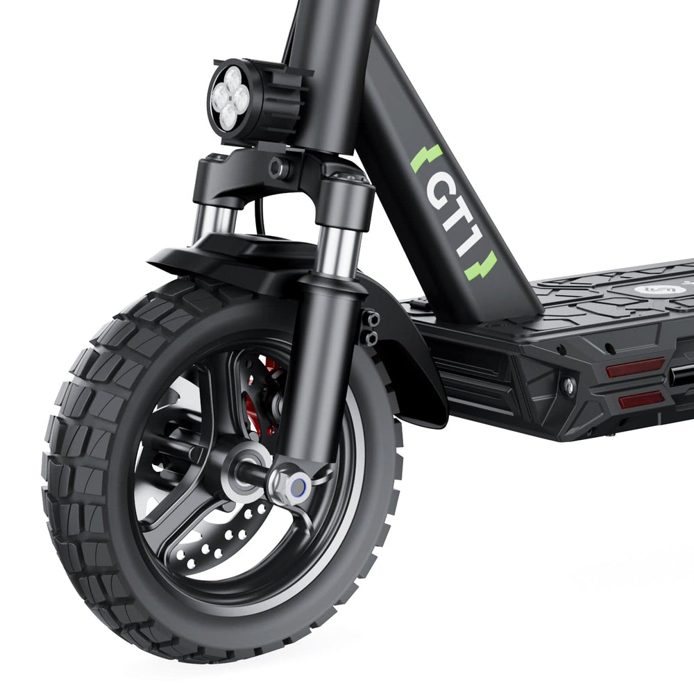 Close-up of an GT1 electric scooter's front wheel featuring a rugged tire, disc brake, and a dual LED headlight mounted above. The frame is marked with "GT1" in green, and the deck has a textured surface for grip.
