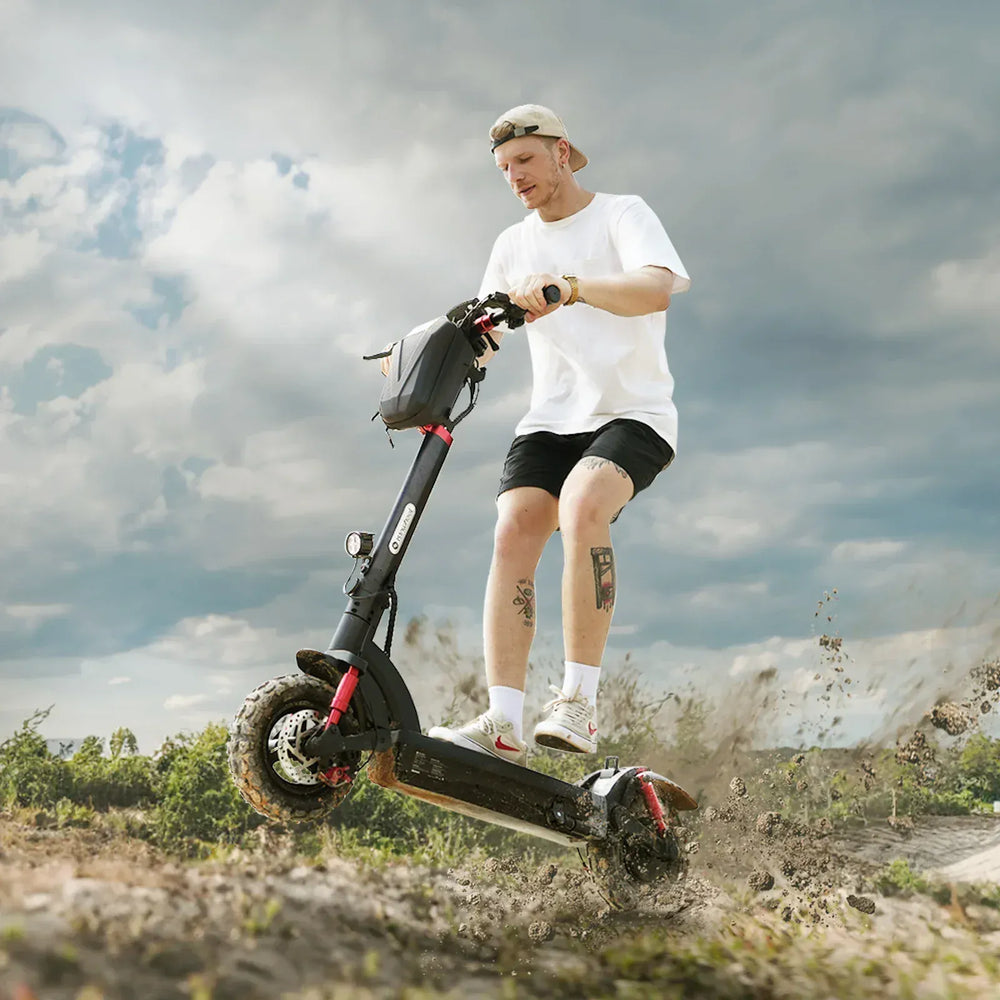 A young man in a white t-shirt, black shorts, and a backwards cap rides the iSinwheel Electric Scooter 1200W All-Terrain Foldable Off-Road E-Scooter – GT2, kicking up dirt as he speeds across rugged ground under a cloudy sky.