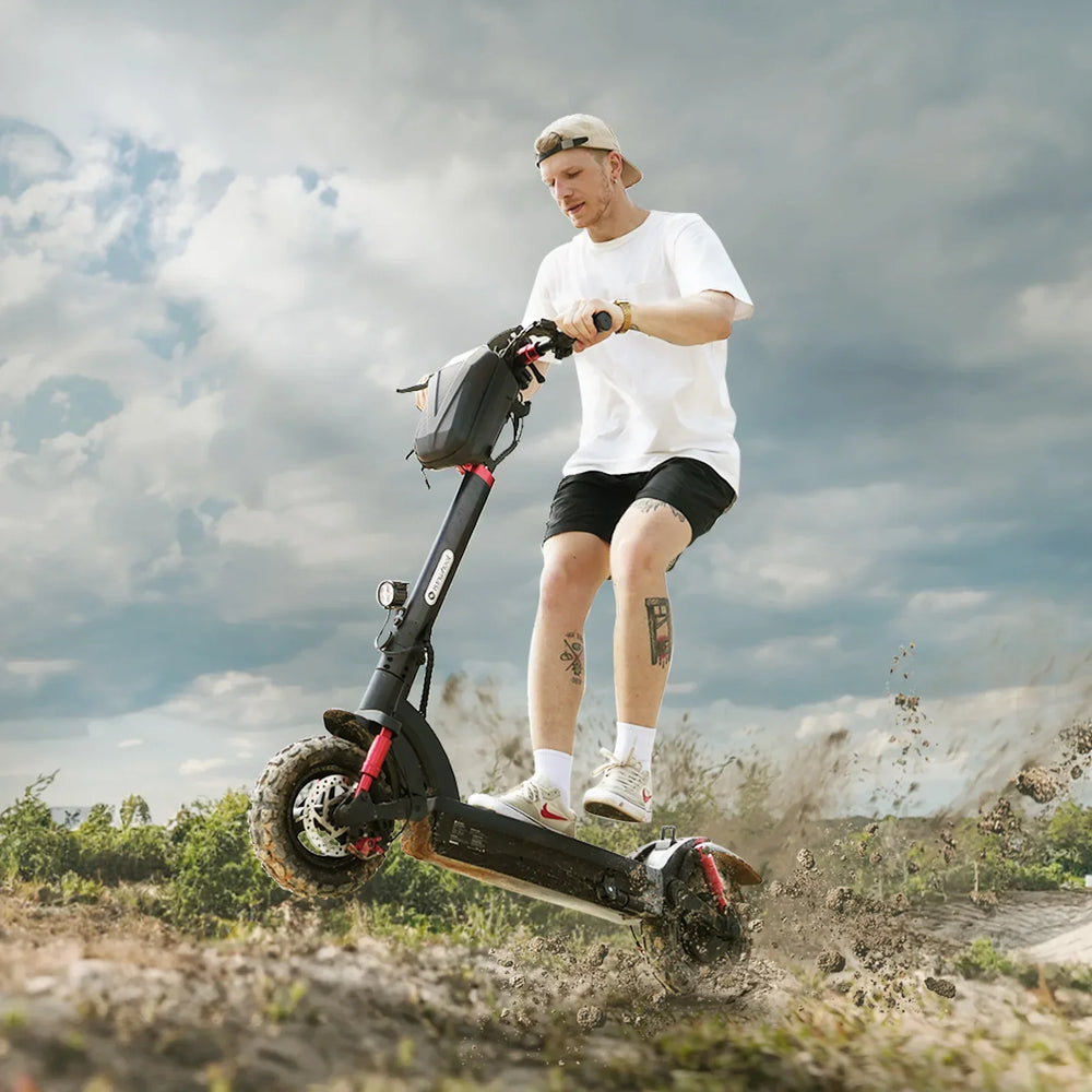 A person rides an electric scooter GT2 off-road, kicking up dirt, under a cloudy sky. They're wearing a white t-shirt, black shorts, and a backwards cap, with visible tattoos on their legs.