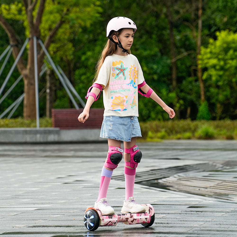 A young girl wearing a helmet and protective gear rides a pink hoverboard H2 on a wet pavement. She is dressed in a colorful t-shirt, a denim skirt, and knee-high socks, surrounded by a lush green park setting.