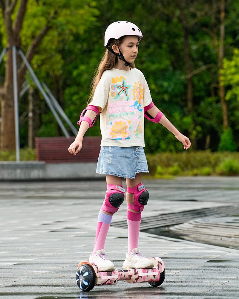 A young girl wearing a helmet and protective gear rides a pink hoverboard H2 on a wet pavement. She is dressed in a colorful t-shirt, a denim skirt, and knee-high socks, surrounded by a lush green park setting.