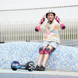 A young person wearing a helmet and pink protective gear sits on a blue tiled surface, adjusting their helmet. A hoverboard H2 rests nearby on the ground. They wear a colorful t-shirt, denim shorts, and black sneakers, with a modern building in the background.