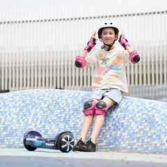 A young person wearing a helmet and pink protective gear sits on a blue tiled surface, adjusting their helmet. A hoverboard H2 rests nearby on the ground. They wear a colorful t-shirt, denim shorts, and black sneakers, with a modern building in the background.