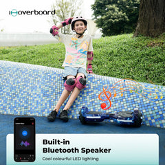 A young person wearing a helmet and pink protective gear sits on a colorful tiled wall, smiling next to a blue hoverboard. The hoverboard H2 features built-in Bluetooth speakers and cool, colorful LED lighting. An app interface is visible on a smartphone in the foreground.