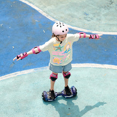 A child wearing a pink helmet and protective gear rides a hoverboard H2 on a blue and green outdoor surface, balancing with arms outstretched. The child is dressed in a colorful t-shirt and denim shorts, enjoying the activity.