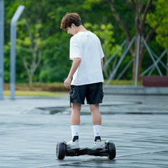 A person wearing a white t-shirt and black shorts is riding a hoverboard H8pro with large wheels on a wet pavement, surrounded by lush green trees.