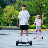 A young man in a white t-shirt and black shorts rides a hoverboard H8pro on a paved path, while a girl in a colorful shirt and protective gear follows behind on another hoverboard. The setting is a lush, green park with trees in the background.