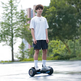 A person wearing a white T-shirt and black shorts is riding a hoverboard H8pro with colorful lights on its wheels, set in an outdoor area with trees and a building in the background.