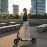 A woman stands on an electric scooter s10max in an urban park, wearing a dark sweater and light pants, with modern buildings and palm trees in the background.