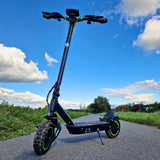 A black electric scooter S10max with green accents stands on a paved path, surrounded by lush greenery and under a bright blue sky with scattered clouds.