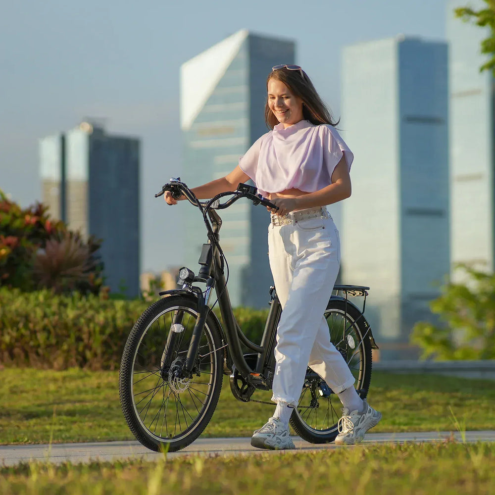 A woman in a light purple top and white pants walks her black bicycle along a grassy path, with modern skyscrapers visible in the background under a clear sky.