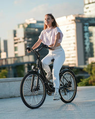 A woman in casual white clothing rides a black bicycle in an urban setting, with modern buildings in the background and sunlight casting a warm glow.