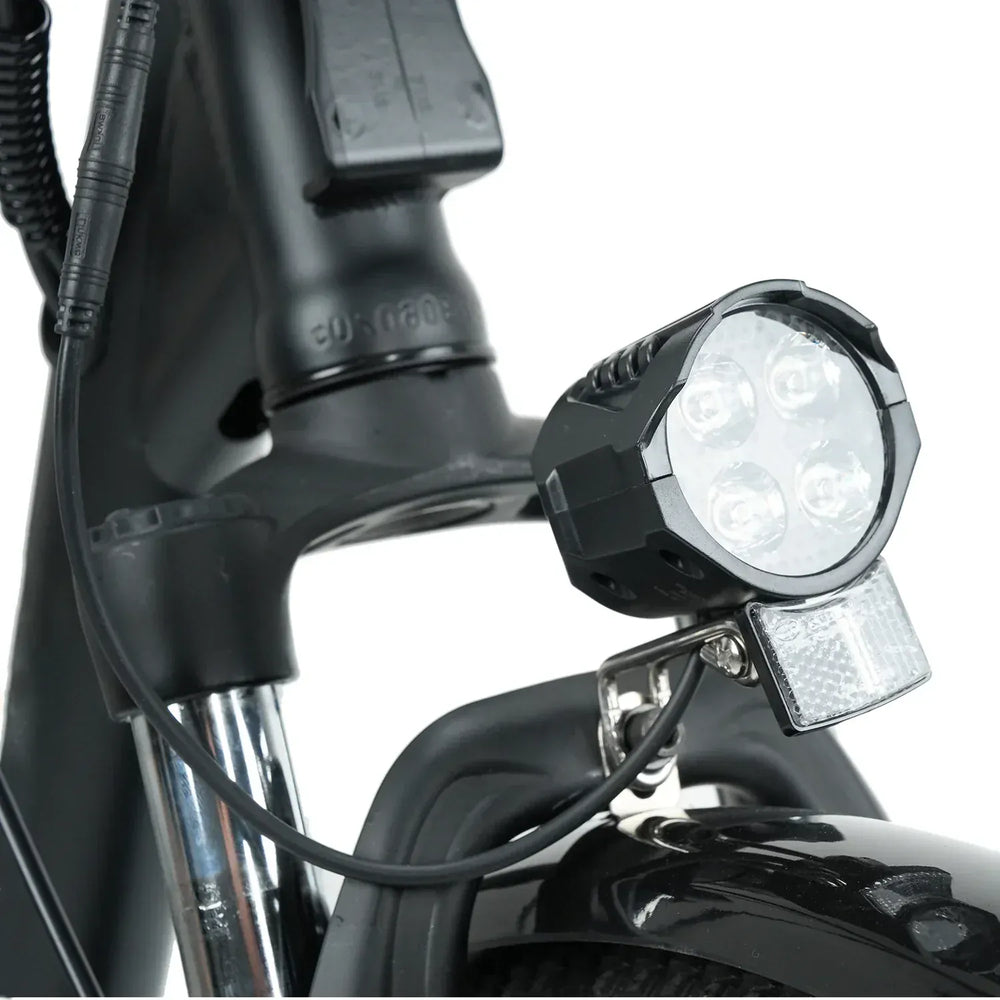A close-up of a bicycle's front section features a bright LED headlight with four bulbs and a reflector mounted above the front wheel on a black frame.