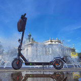 A sleek black electric scooter e9pro stands in front of a lively fountain, with water jets creating a dynamic backdrop. The scooter's reflection is visible on the wet pavement, and a historic building with ornate details is in the background under a clear blue sky.