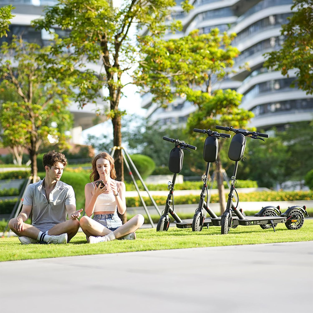 A young man and woman sit on the grass in a park, engaged with a smartphone, while three e9pro electric scooters are parked nearby. Behind them, lush trees and modern buildings create a lively urban backdrop.