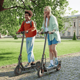 Two people ride electric scooters i8 along a cobblestone path in a park, surrounded by lush greenery. They are smiling and enjoying the sunny day, with one wearing a backpack and headphones, and the other in a light green shirt and denim shorts.