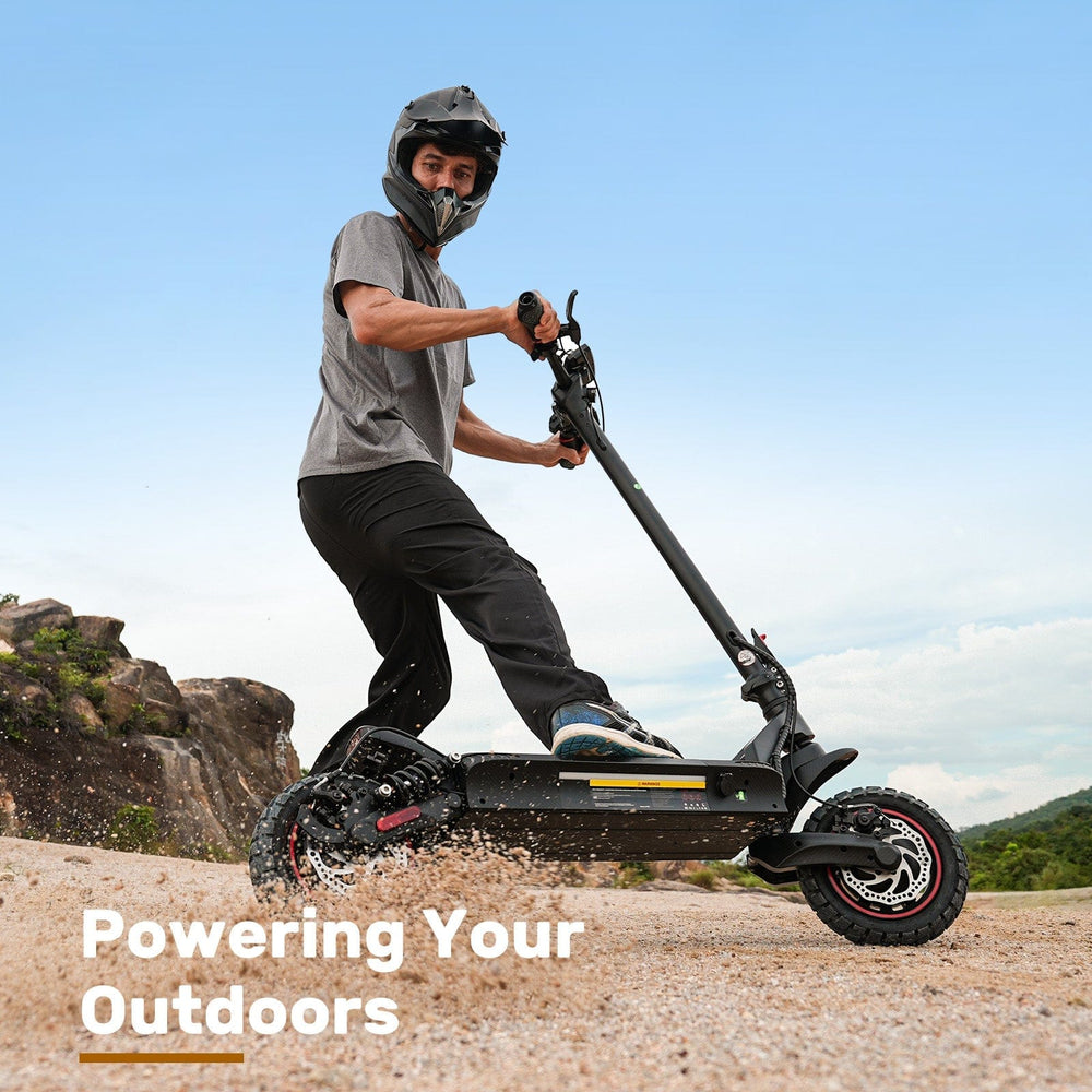 A person wearing a helmet rides an electric scooter ix7pro across sandy terrain, kicking up dust, against a backdrop of blue sky and rocky hills.