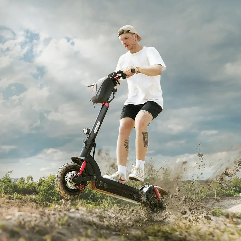 A person rides an electric scooter GT2 off-road, kicking up dirt, under a cloudy sky. They're wearing a white t-shirt, black shorts, and a backwards cap, with visible tattoos on their legs.