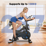 A woman in workout attire is using a stationary exercise bike QK116C in a bright, modern living room. The bike is black with blue accents, and text on the image highlights its weight capacity of 130 kg (287 lbs).