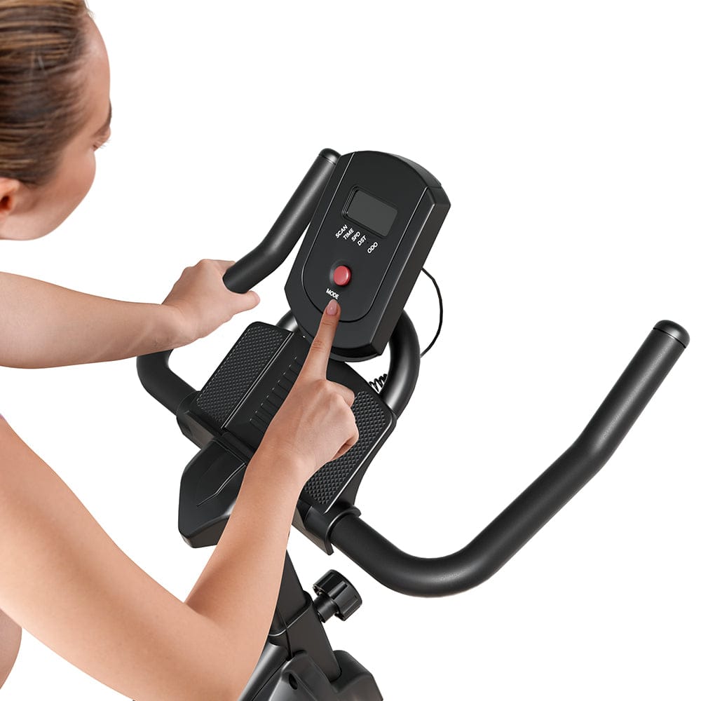 A person is using a stationary exercise bike QK116C, pressing a button on the digital display. The bike has black handlebars and a textured footrest, with a clear focus on the control panel.