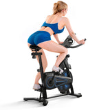 A woman in blue athletic wear is using a stationary exercise bike QK116C, positioned in an indoor setting. She is leaning forward, gripping the handlebars, and wearing white sneakers. The bike features a sleek design with blue accents.