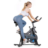 A woman in a white crop top and blue leggings is exercising on a stationary bike QK116C. She is leaning forward, gripping the handlebars, with her hair tied back in a ponytail. The bike features a black and blue design, and she is wearing white sneakers.