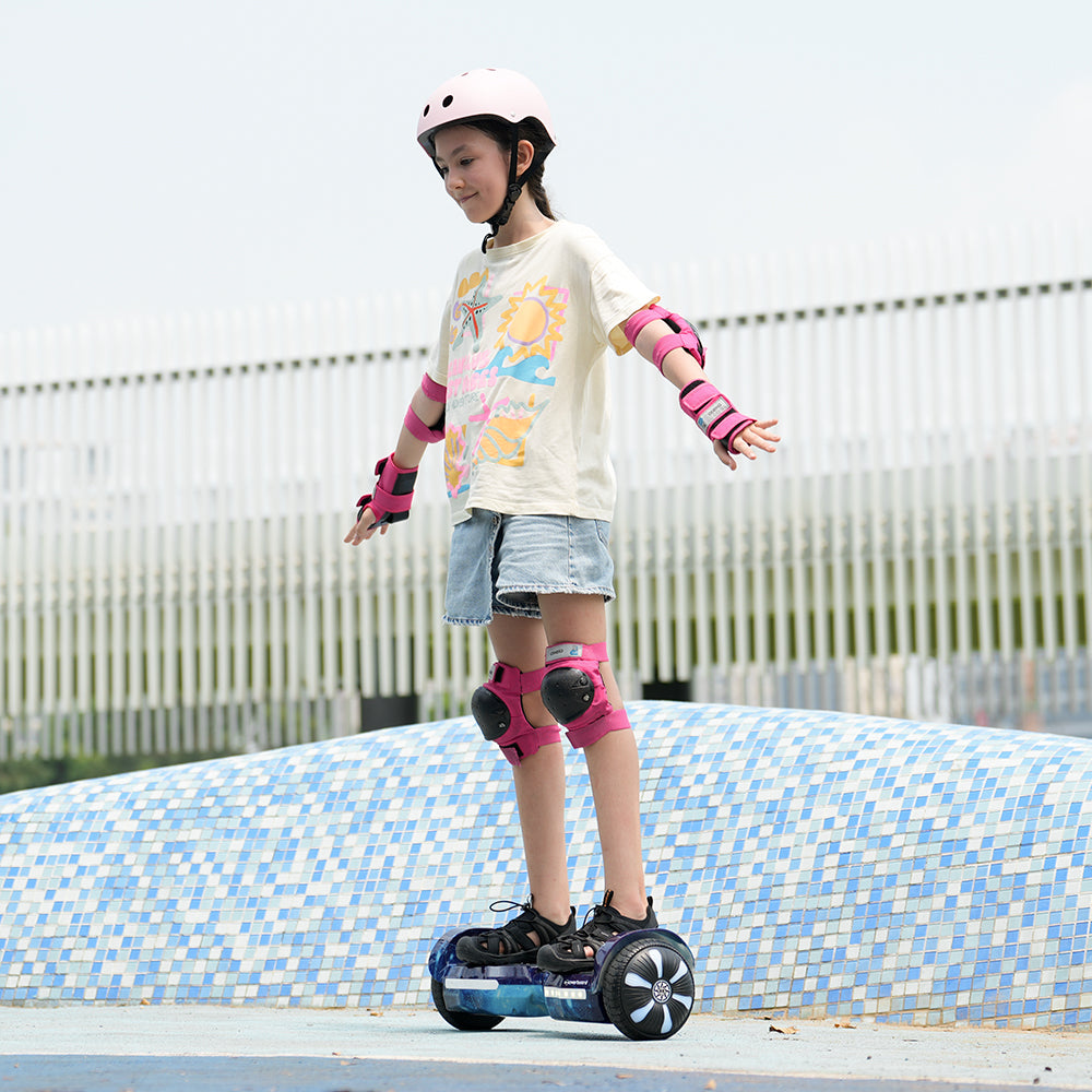 A young girl wearing a helmet and pads rides the iHoverboard Hoverboard for Kids Aged 6+ (6.5"-H2) with Bluetooth and LED, balancing with arms outstretched as she enjoys her outdoor ride on a tiled blue surface.