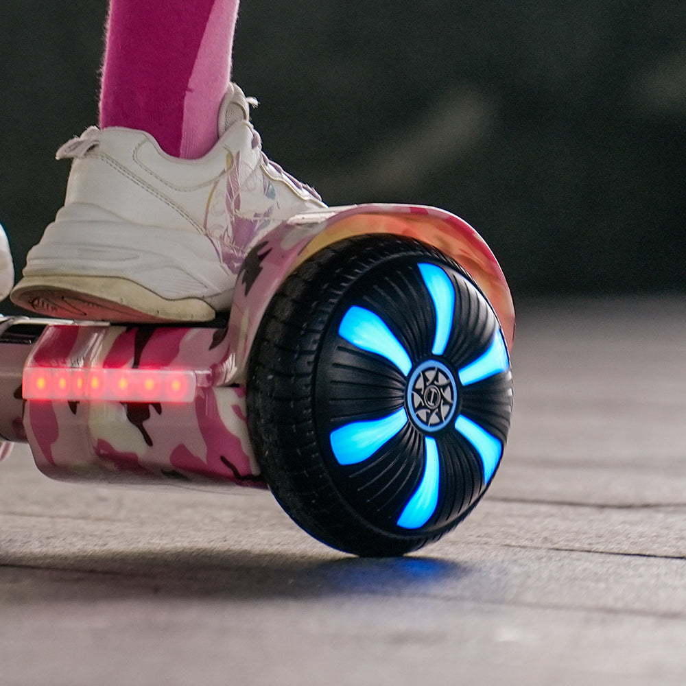 Close-up of a person in white sneakers and pink leggings riding the iHoverboard Hoverboard for Kids Aged 6+, a self-balancing hoverboard with Bluetooth, blue LED lights on the 6.5" wheels, and a pink camouflage design.