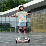 A young girl in a t-shirt and shorts balances with outstretched arms on the iHoverboard Hoverboard for Kids Aged 6+, a 6.5" self-balancing scooter with Bluetooth and LED, wearing a helmet and pads for safety outside a modern building.