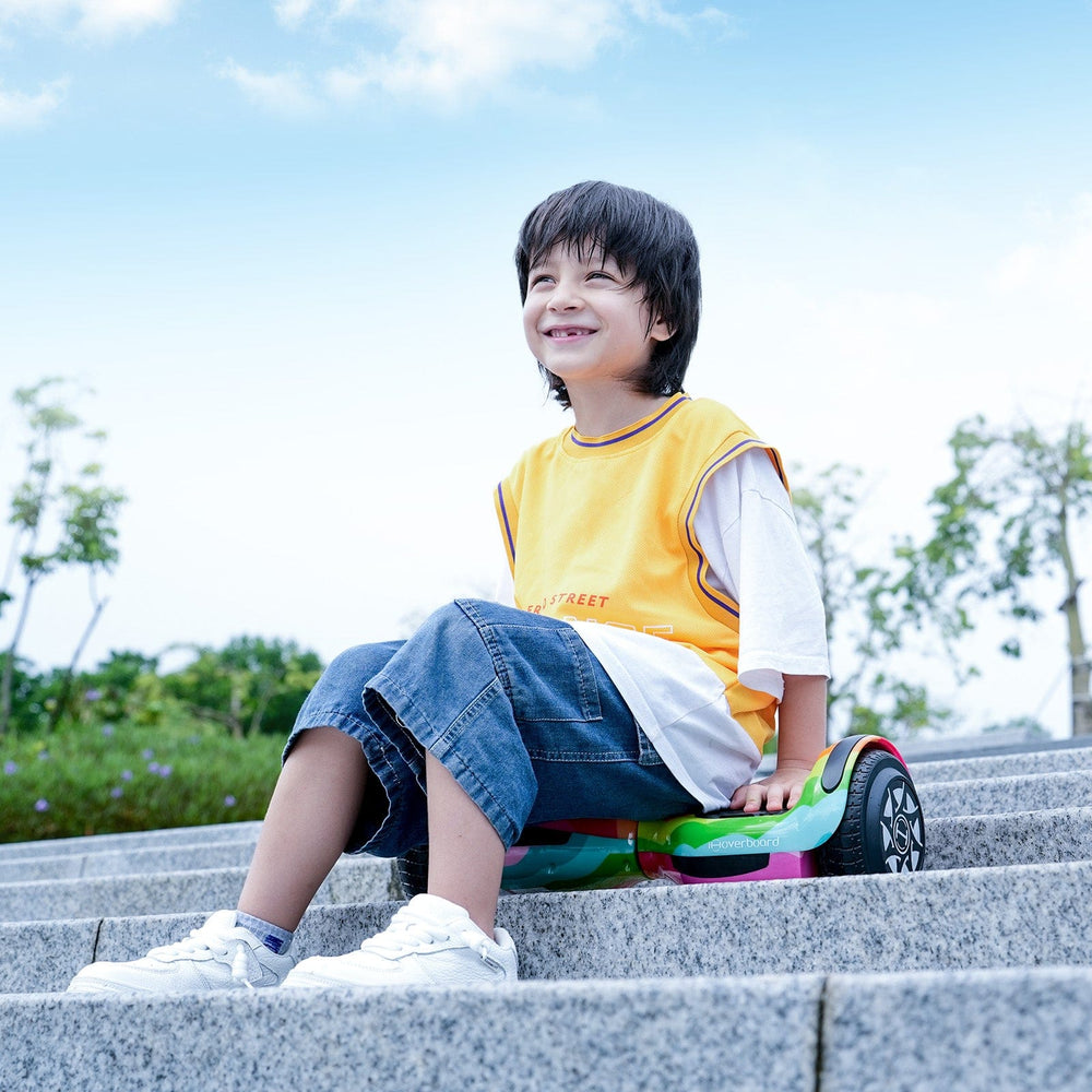 A smiling boy in a yellow shirt and denim shorts sits on the iHoverboard Hoverboard for Kids, 6.5" Self-Balancing with Bluetooth LED Lights (CE Certified-H1), at the top of stone steps under a bright blue sky.
