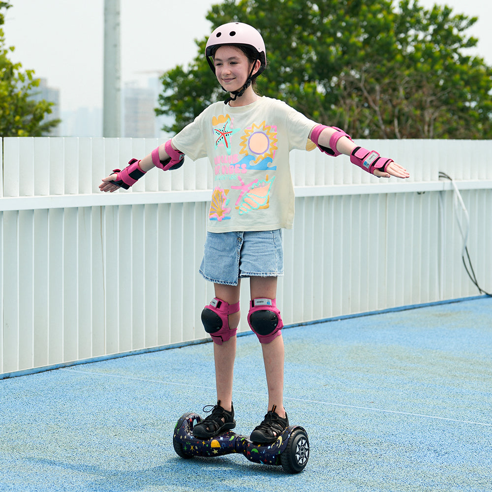 A smiling child in a helmet and pink gear rides the iHoverboard Hoverboard for Kids Aged 6+ (6.5"-H2) with Bluetooth & LED, balancing on a blue outdoor surface, trees and city skyline in the background.
