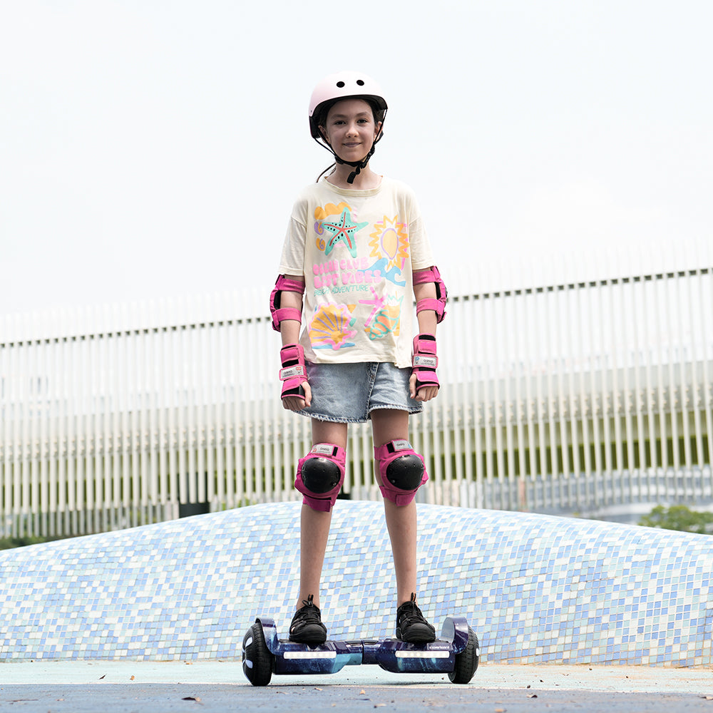 A young girl in a helmet and pink gear stands on the iHoverboard Hoverboard for Kids Aged 6+, featuring Bluetooth and LED lights, outdoors. She wears a colorful t-shirt and denim shorts, with a mosaic-tiled surface and modern fence behind her.