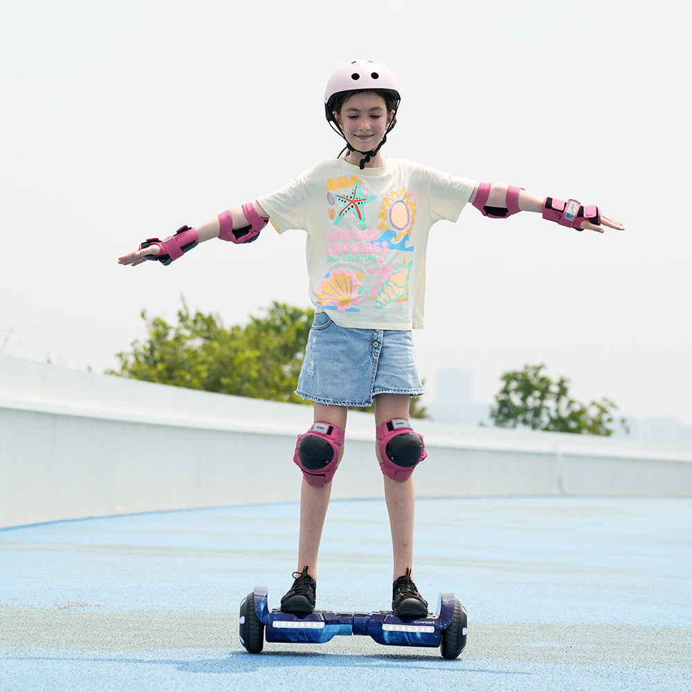 A young girl wearing safety gear rides the iHoverboard Hoverboard for Kids Aged 6+, Self Balancing Hoverboard with Bluetooth & LED 6.5"-H2, on a blue outdoor track, arms outstretched. Trees and a cityscape appear in the background.