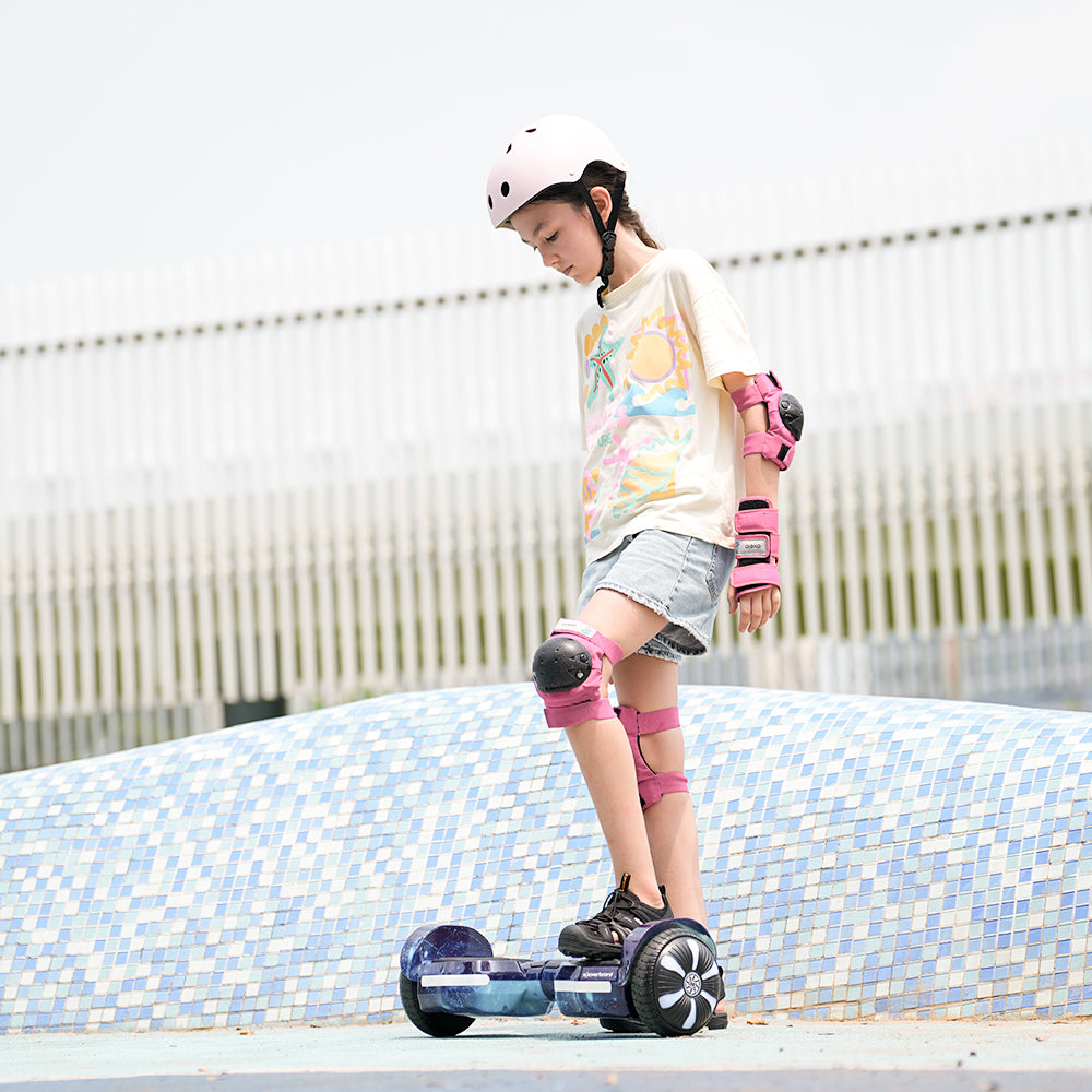 A young girl in a helmet and pads rides the iHoverboard Hoverboard for Kids Aged 6+, a self-balancing hoverboard with Bluetooth and LED 6.5"-H2, at a skate park. She wears light clothes and stands on a blue-tiled surface.