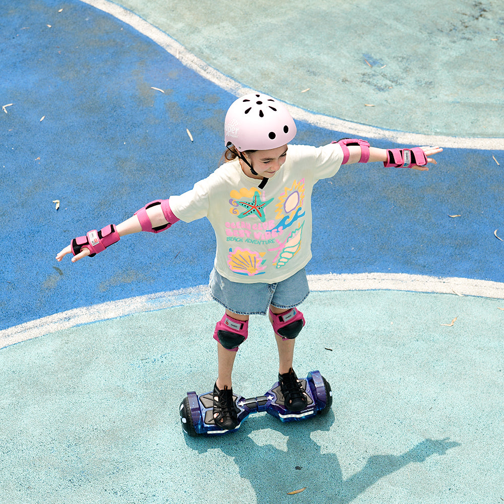 A young child in a pink helmet and pads rides the iHoverboard Hoverboard for Kids Aged 6+, a self-balancing Bluetooth model with LED 6.5"-H2, on a blue and teal outdoor sports court, arms outstretched for balance.