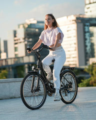 A woman in casual white clothing rides a black bicycle in an urban setting, with modern buildings in the background and sunlight casting a warm glow.