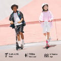 Two children ride scooters ik1s on a sunny day, both wearing helmets for safety. The child on the left rides a black scooter, dressed in a black and white outfit, while the child on the right rides a pink scooter, wearing a pink jacket and white shorts. A pastel pink wall serves as the backdrop.