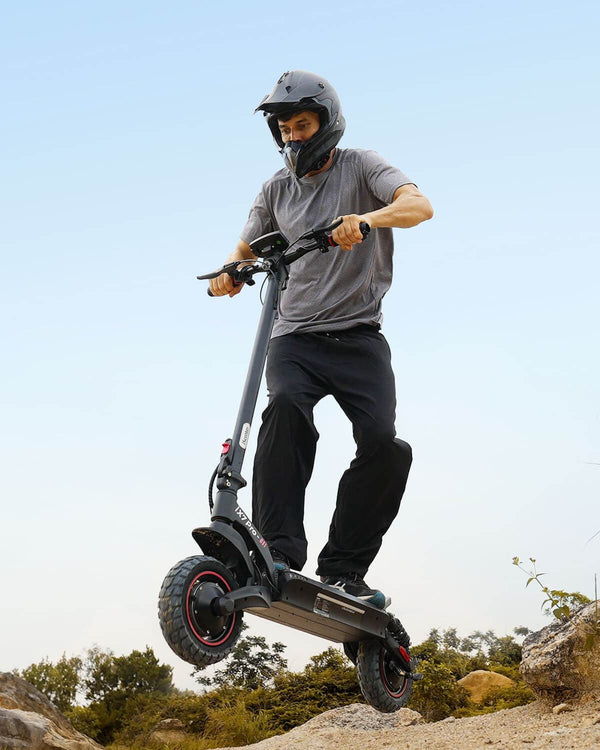 A young girl wearing a pink helmet, elbow and knee pads, and a colorful shirt is balancing with arms outstretched on a pink camouflage hoverboard. She stands on a smooth, wet pavement in front of a modern glass building, looking focused and confident.