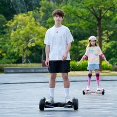 A young man in a white t-shirt and black shorts rides a hoverboard H8pro on a paved path, while a girl in a colorful shirt and protective gear follows behind on another hoverboard. The setting is a lush, green park with trees in the background.