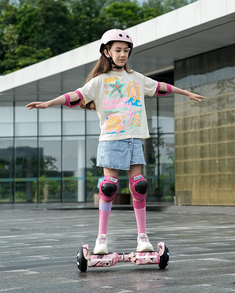 A young girl wearing a pink helmet and protective gear is balancing on a pink hoverboard H2 outside a modern building. She has her arms outstretched for balance, and is dressed in a colorful t-shirt and denim skirt.