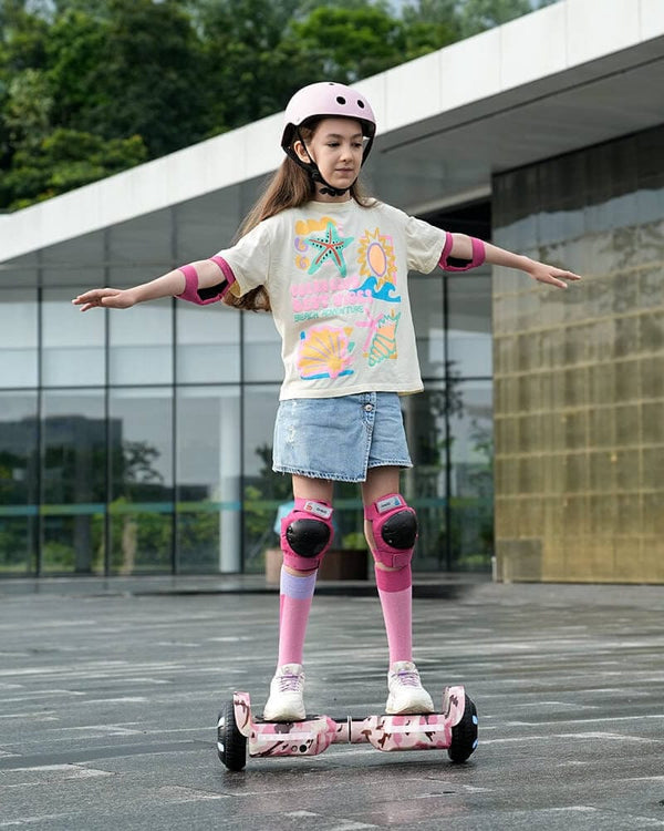 A young girl balances confidently on a pink camouflage hoverboard, wearing a helmet, knee pads, and elbow pads for safety. She sports a colorful T-shirt with a summer-themed design, a denim skirt, and knee-high socks, standing in front of a modern glass building.