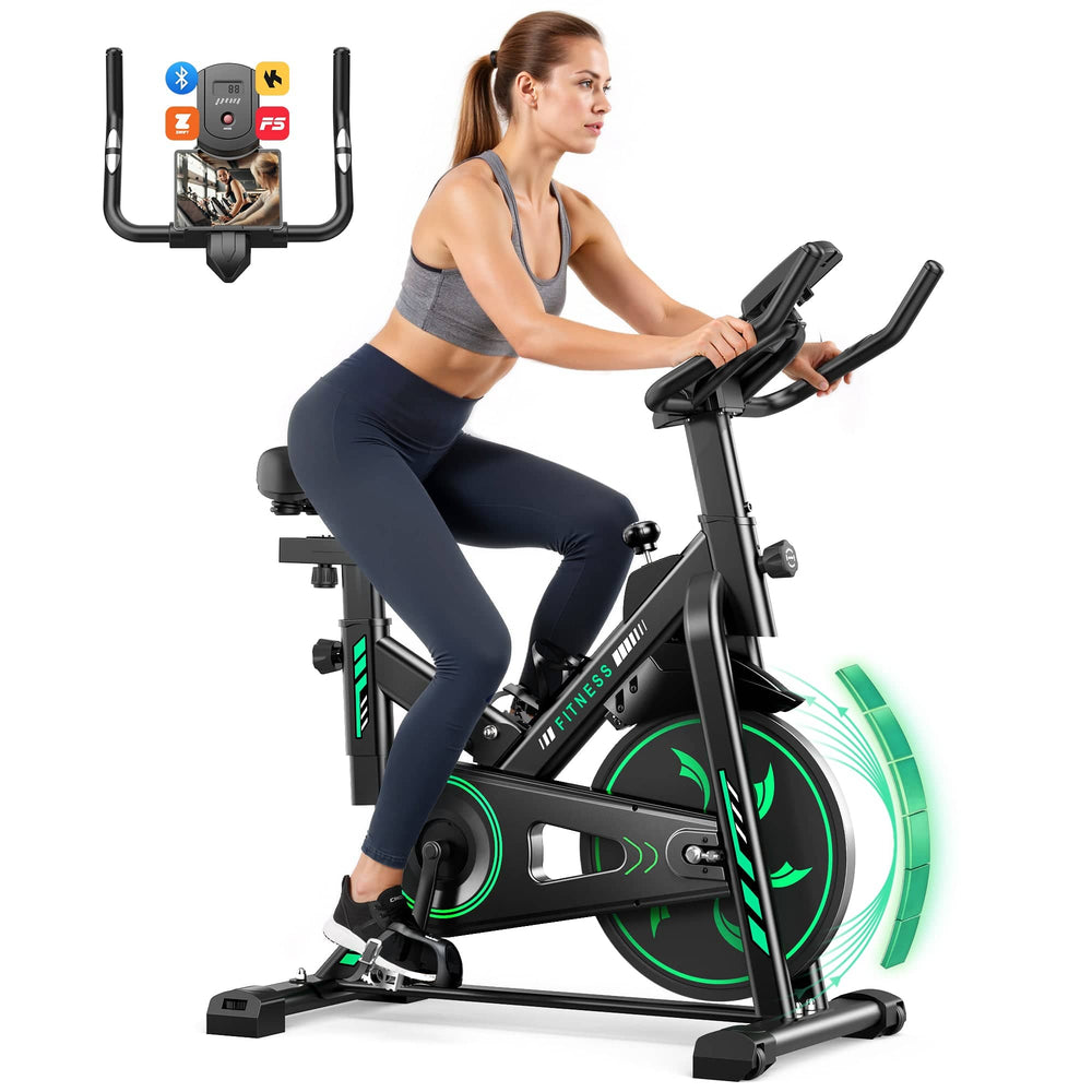 A woman is exercising on a sleek, black stationary bike with green accents. She is wearing a gray sports bra and navy leggings, focused on the digital display. The bike features a high-tech console with Bluetooth and app connectivity symbols.