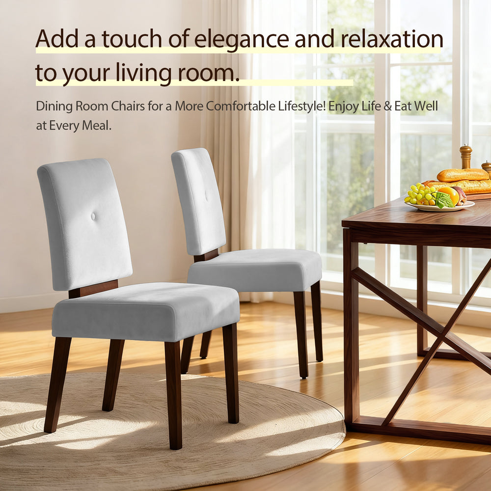 Two elegant gray dining chairs with wooden legs are placed on a circular rug in a sunlit room, next to a wooden table topped with fruit and bread.