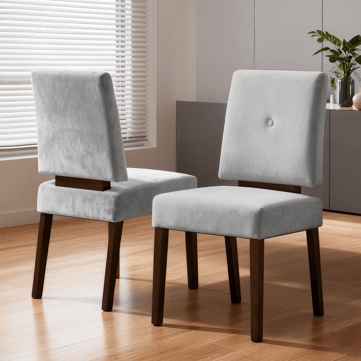 Two modern chairs with light gray upholstery and dark wooden legs are positioned on a wooden floor in a sunlit room, with a plant and cabinet visible in the background.