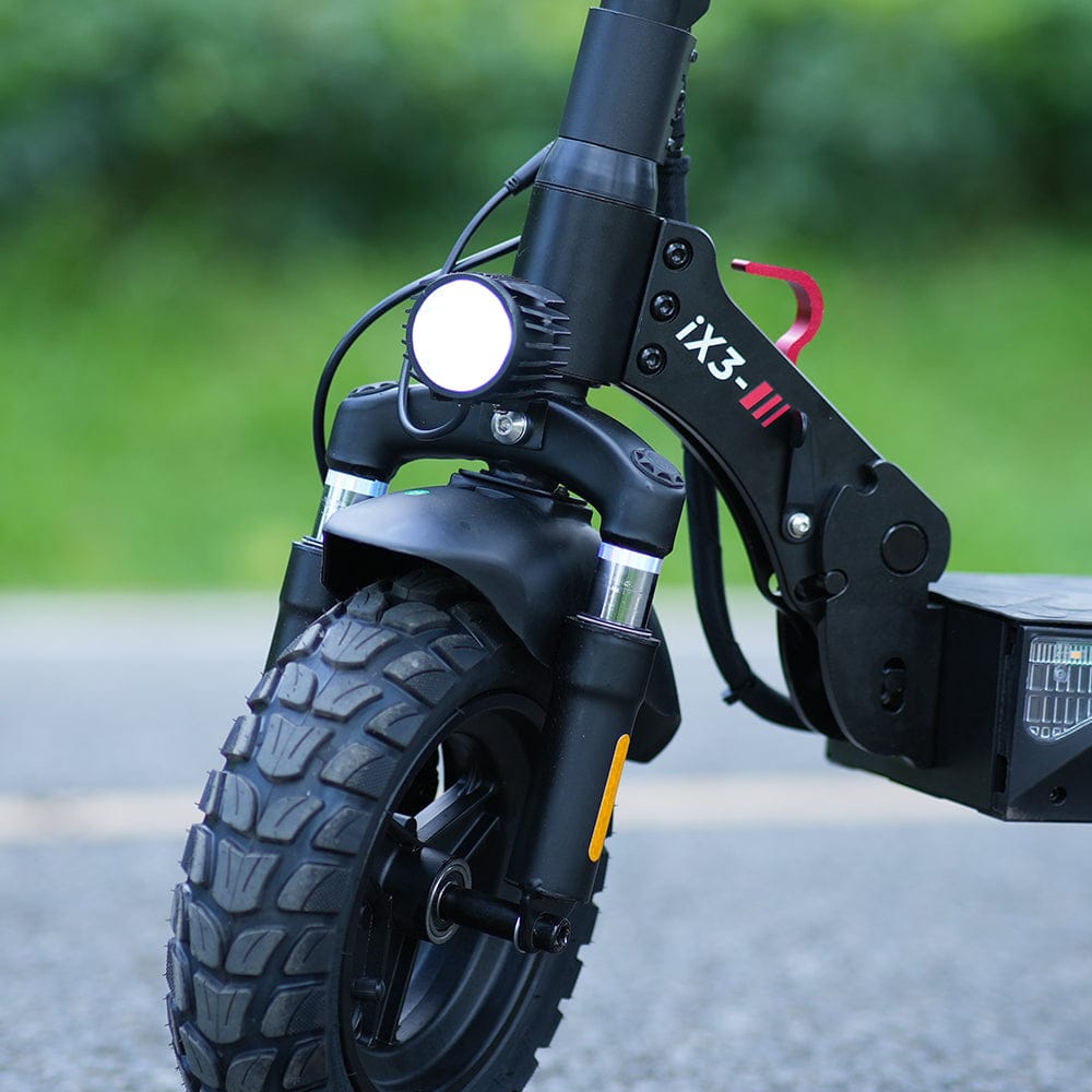 Close-up of the front wheel of a black electric scooter ix3 with rugged tires, featuring a bright headlight and visible branding "iX3" on the frame, set against a blurred green background.